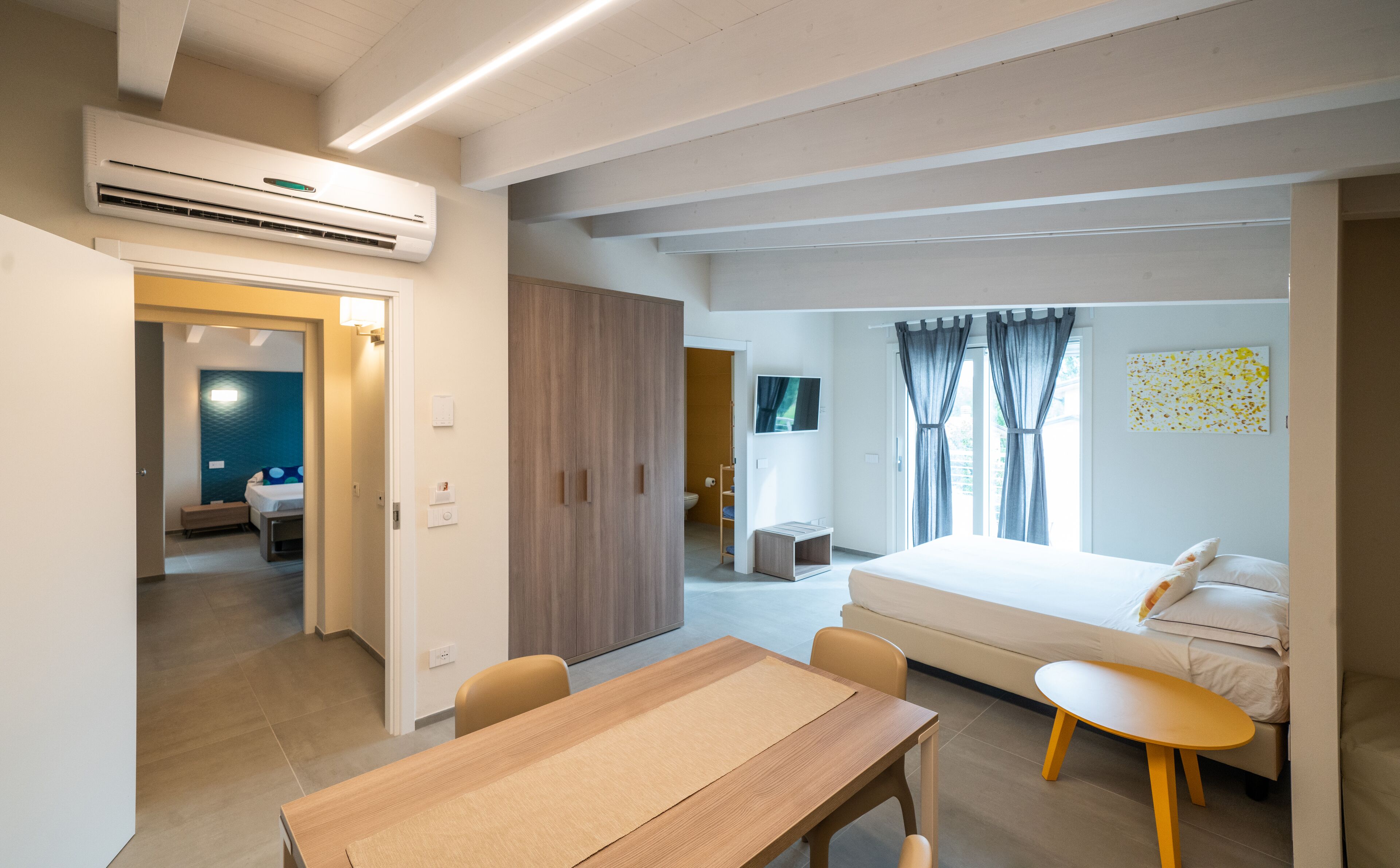 A modern hotel room with a wooden table, a bed, a TV, and a bathroom visible through an open door.