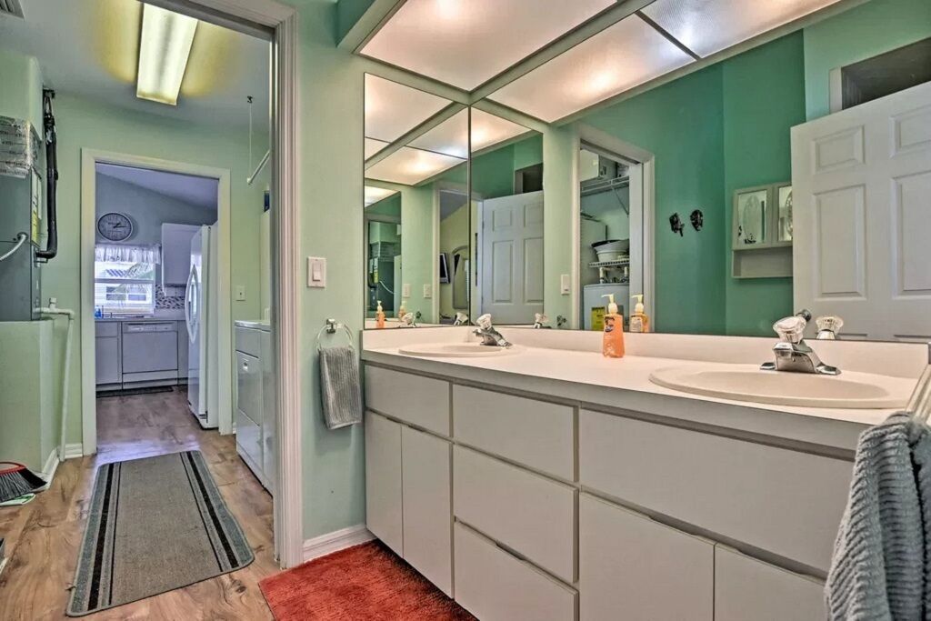 A bathroom with a large mirror, two sinks, and a door leading to a kitchen.