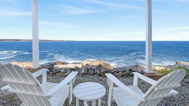 Un área de estar al aire libre con techo, con sillas Adirondack blancas y una pequeña mesa, con vista a un litoral rocoso y al océano.