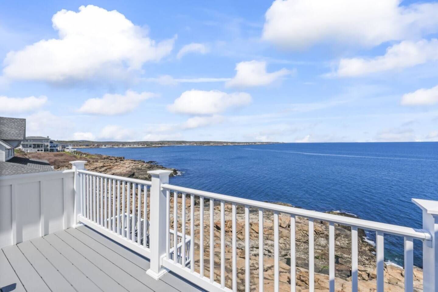 A coastal property with a wooden deck and white railing, overlooking a rocky shoreline and the sea.