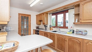 A kitchen with wooden cabinets, a white countertop, and a window with curtains. - Simdde Wen -  a bungalow that sleeps 6 guests  in 3 bedrooms (Beaumaris)