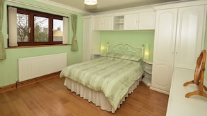 A bedroom with a bed, a radiator, a window with curtains, and white cabinets. - Simdde Wen -  a bungalow that sleeps 6 guests  in 3 bedrooms (Beaumaris)