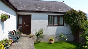 A small house with a dark brown door, a window with brown shutters, and a small garden with potted plants and a bench. - Simdde Wen -  a bungalow that sleeps 6 guests  in 3 bedrooms (Beaumaris)