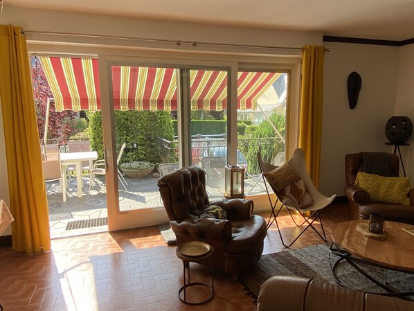 A living room with a brown leather sofa, a white armchair, and a wooden coffee table. There is a sliding glass door leading to an outdoor area with a striped awning.