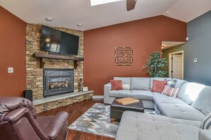 A living room with a stone fireplace, a grey sectional sofa, a brown leather chair, and a flat-screen TV mounted on the wall. - East AVL, 7 miles to Downtown, Game Room, Hot Tub (Asheville)