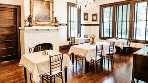 Un espace repas avec des tables et des chaises en bois, un lustre et une cheminée surmontée d’un grand miroir décoratif.