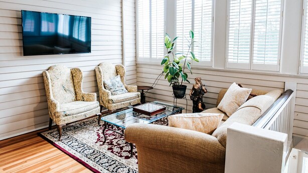 Un salon comprenant deux fauteuils, un canapé, une table basse, un téléviseur et une grande fenêtre avec des volets.