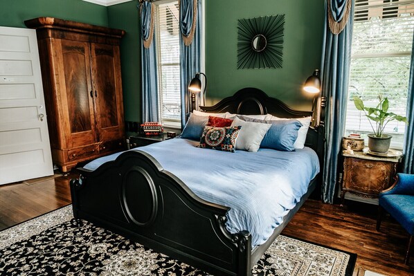Un dormitorio con un armario de madera grande, una cama con muchas almohadas, un espejo decorativo y una planta en maceta cerca de la ventana.