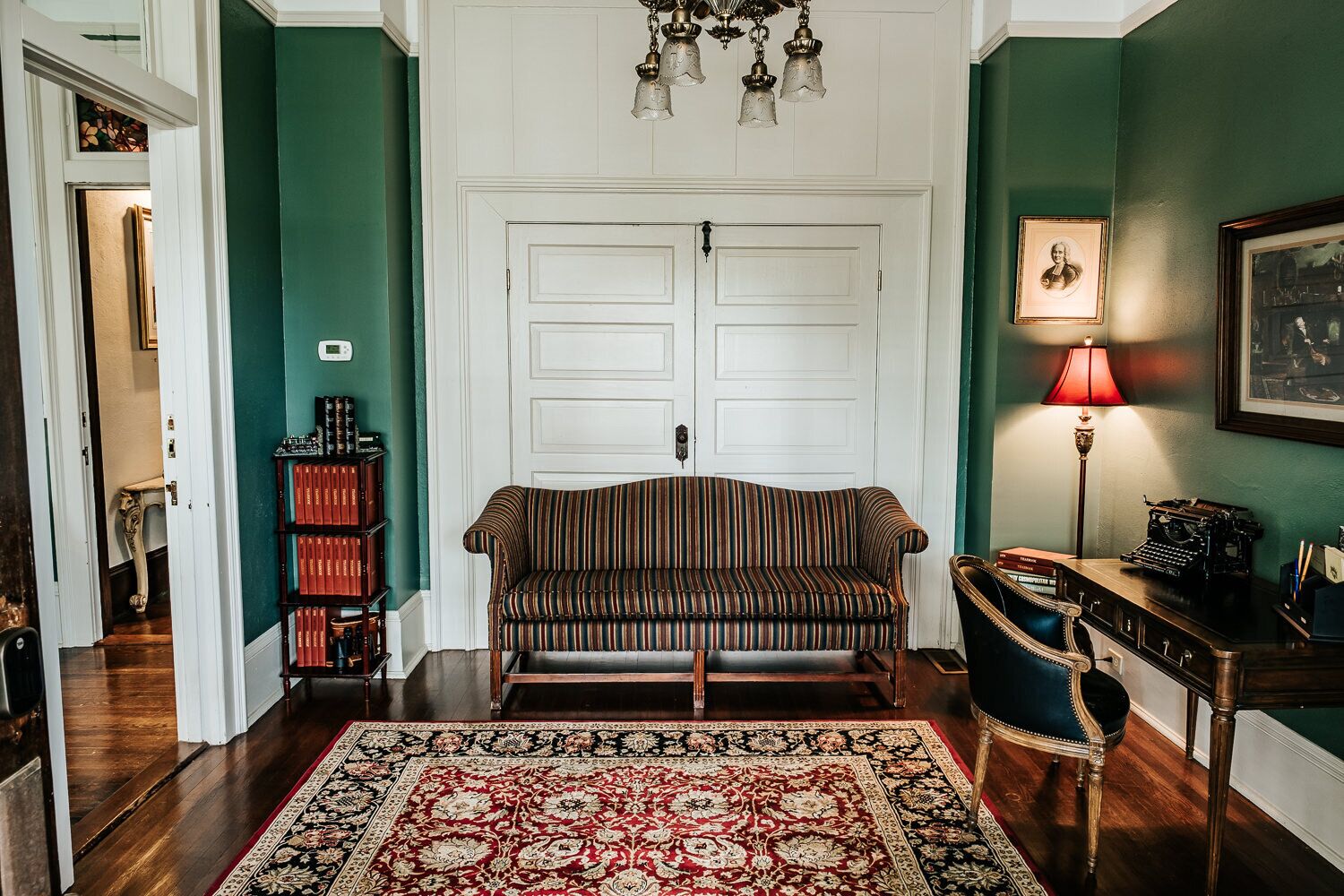 A room with a striped sofa, a red rug, a desk with a typewriter, and a bookshelf.
