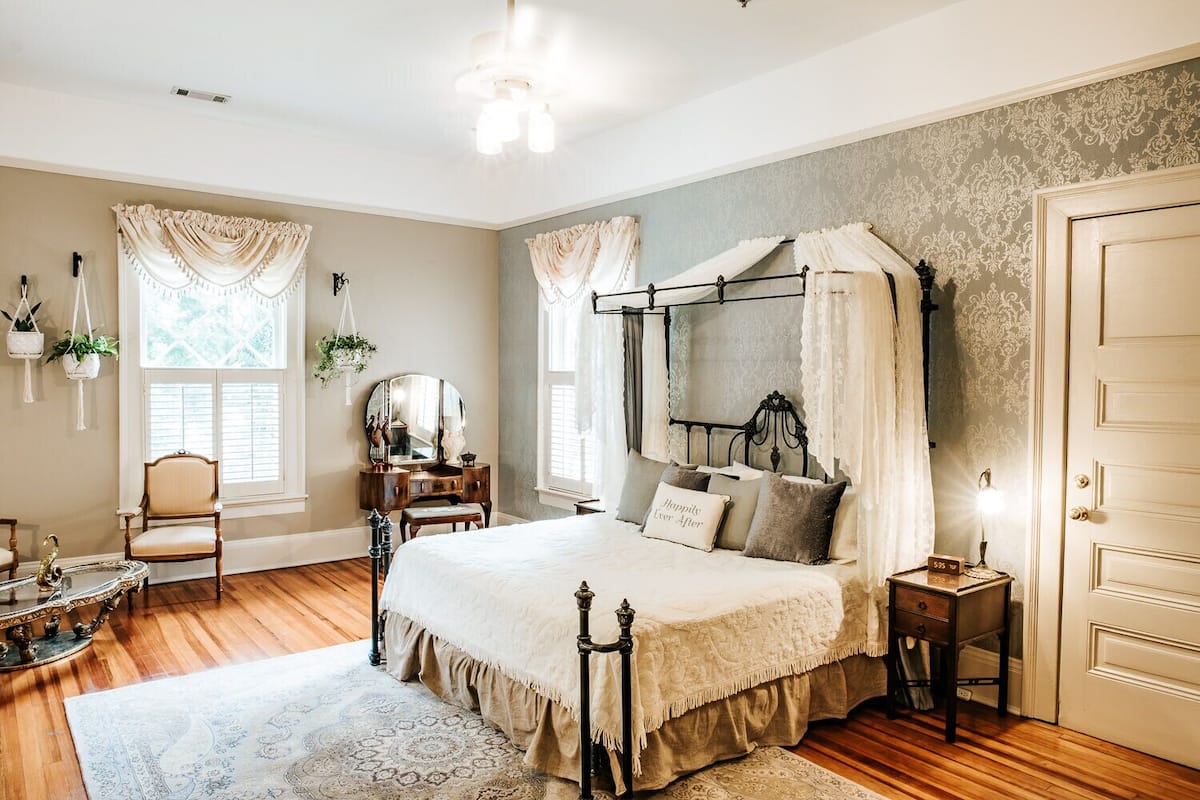 A bedroom with a four-poster bed, a vanity with a mirror, a chair, and a small table with a lamp.