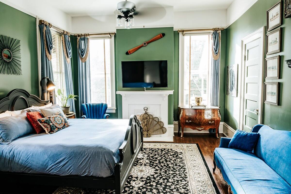 A bedroom with a bed, a blue armchair, a wooden dresser, a television, and a large window with curtains.