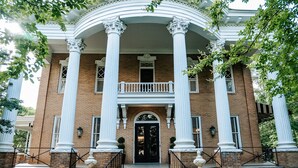 Un grand bâtiment en briques, doté d’une entrée imposante, avec une porte centrale flanquée de deux rangées de colonnes, un balcon et un escalier menant à l’entrée.