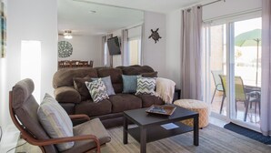 Een woonkamer met een bruine bank, een houten fauteuil en een bijzettafeltje. Er is een groot raam met uitzicht op tuinmeubilair.