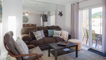 A living room with a brown sofa, a wooden armchair, and a small table. There is a large window with a view of outdoor furniture.