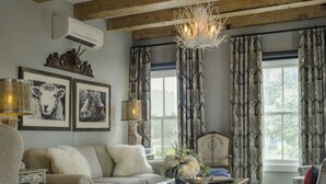 Una sala de estar acogedora con un sofá, un sillón y una mesita de centro. Un candelabro cuelga del techo, y hay cortinas en las ventanas.
