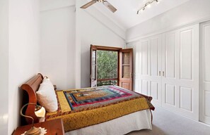 Ein Schlafzimmer mit einem Bett, einem Nachttisch, einer Lampe, einem Fenster mit Blick auf Bäume und einem Schrank mit weißen Türen.
