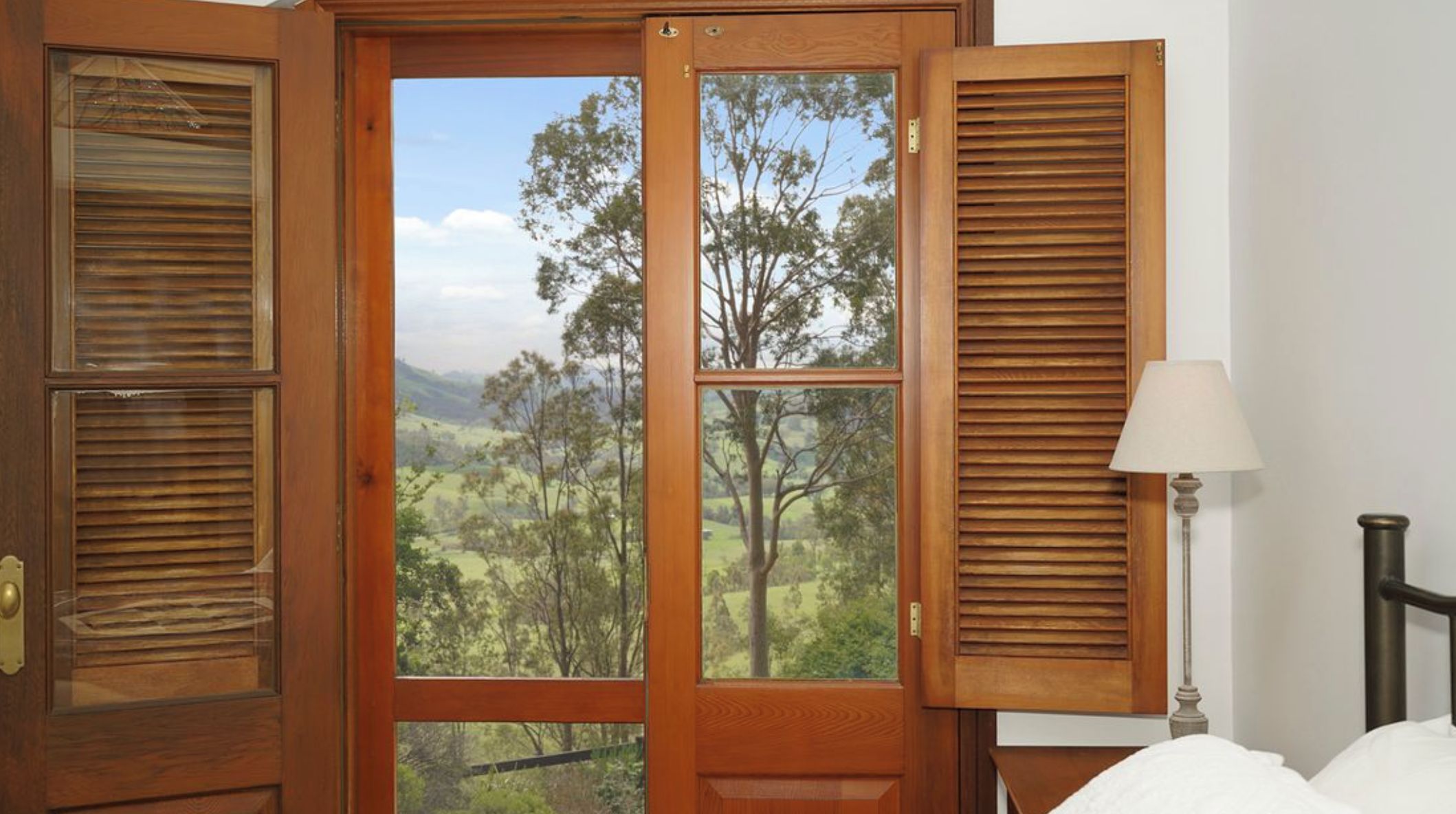 A wooden door with shutters, a view of a landscape with trees and hills, a bedside lamp, and a bed.