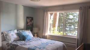 A bedroom with a bed, a nightstand, a lamp, a crib with a teddy bear, and a window with curtains. - Lake Whatcom Summer home, 100' private beach, paddle boards and kayaks! (Bellingham)