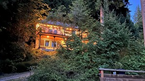 A cabin with a porch and a deck, surrounded by dense forest. - Lake Whatcom Summer home, 100' private beach, paddle boards and kayaks! (Bellingham)