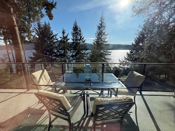 Une table en verre, entourée de chaises, sur un balcon donnant sur un lac, le tout bordé d’arbres.
