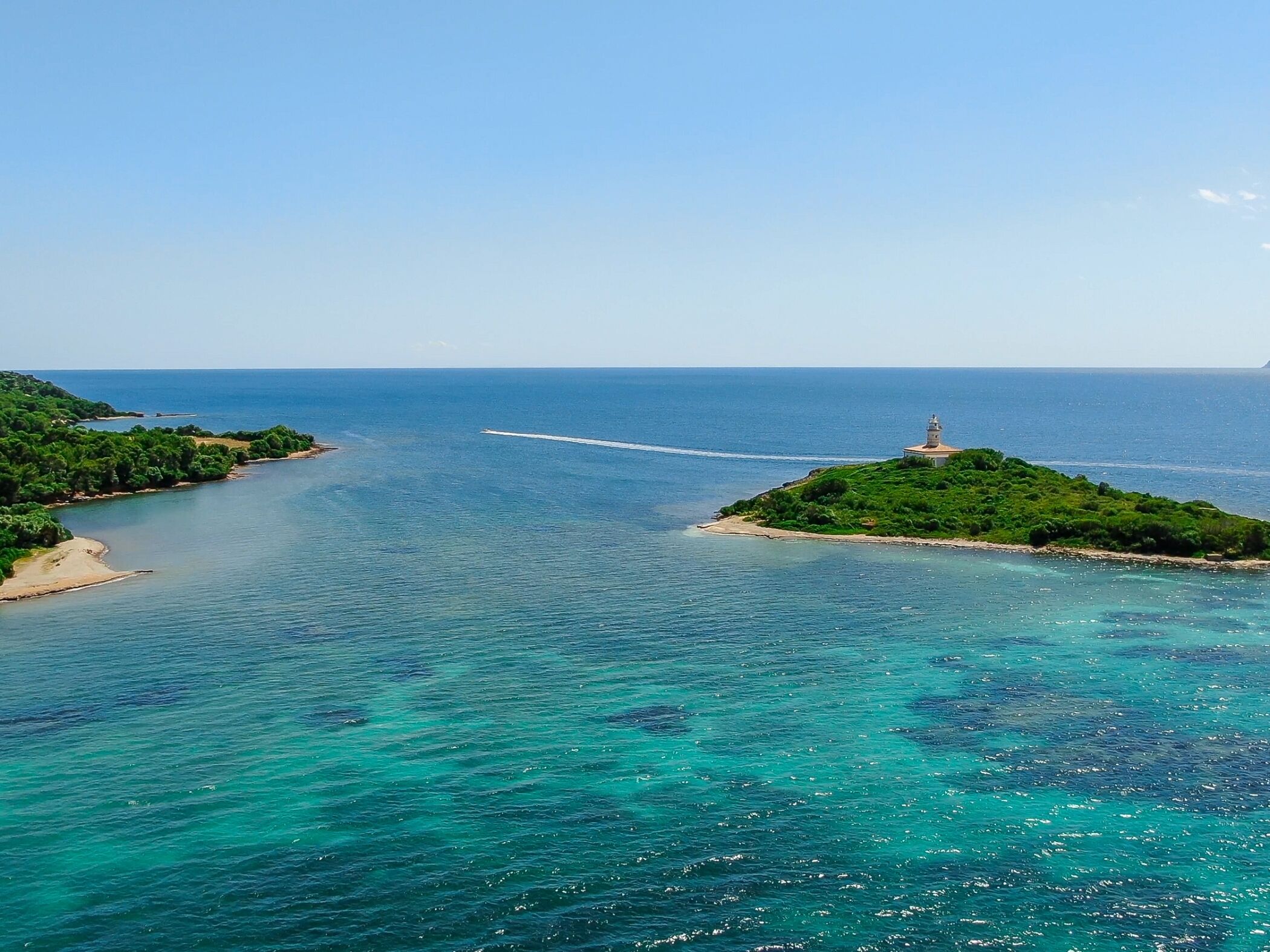 Un paysage côtier avec un phare situé sur une petite île, des eaux d’un bleu limpide et une plage de sable.