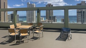 A rooftop terrace with a table and chairs, large windows offering ocean views, and a metal chair. - Ocean Views, Large Balcony + Walk to Everything (Honolulu)