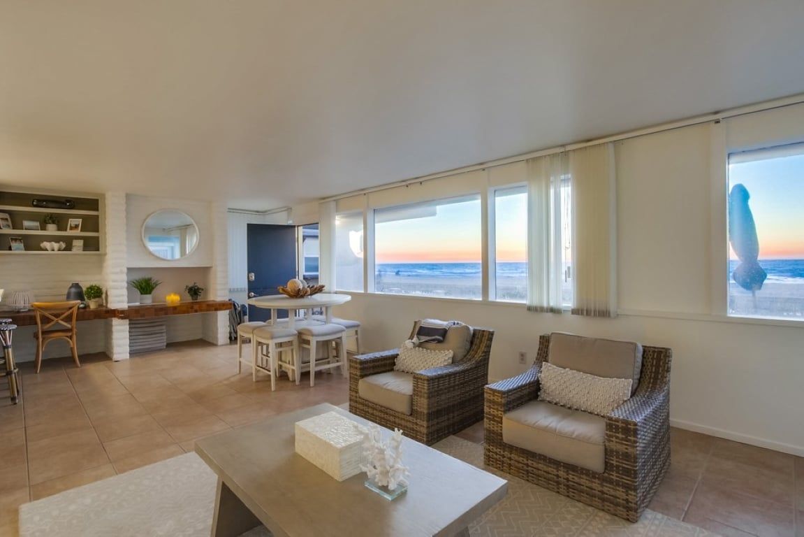 A spacious living area with a view of the sea, a dining table, wicker furniture, and a kitchen area with shelves.