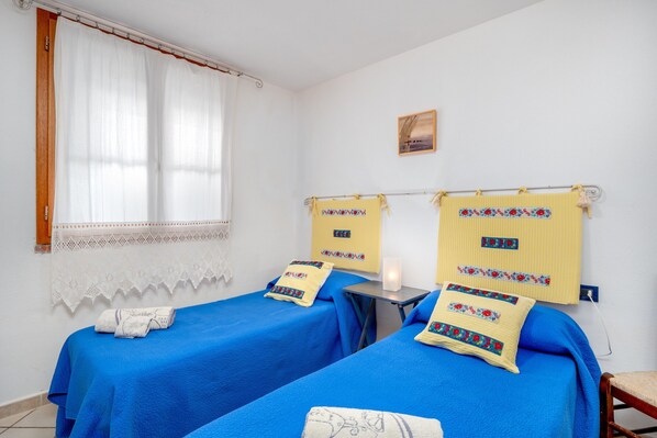 Une chambre avec deux lits, chacun recouvert d’une couvre-lit bleue et agrémenté d’un coussin décoratif. On y trouve une petite table avec une lampe, une fenêtre avec des rideaux blancs et un luminaire fixé au mur.