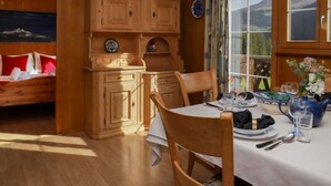 Una habitación acogedora con paredes de madera y un ventanal grande. Hay una mesa de comedor dispuesta con platos, vasos y un jarrón de flores. Se ve una cama con almohadones rojos en una esquina.