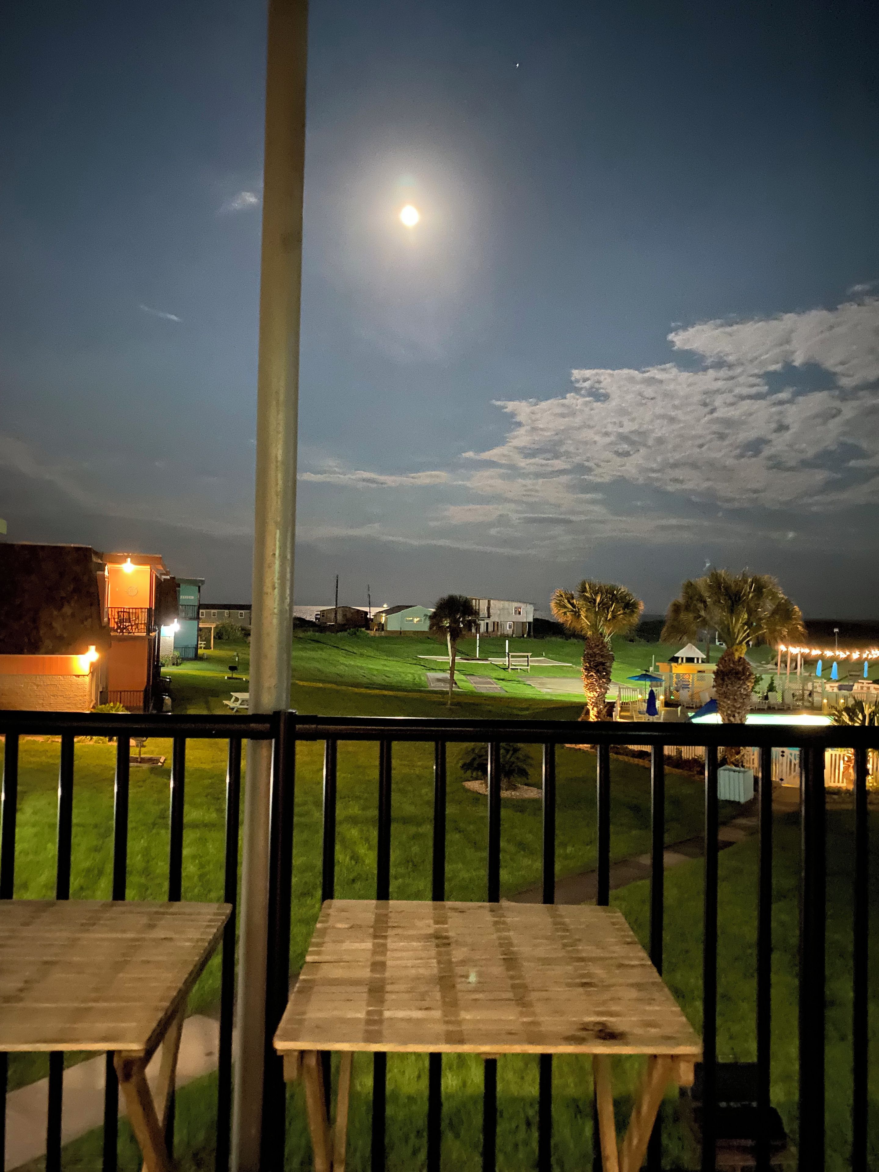 Vista nocturna desde un balcón con una mesa y sillas, una zona ajardinada y un edificio al fondo.