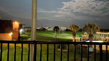 Vista nocturna desde un balcón con una mesa y sillas, una zona ajardinada y un edificio al fondo.
