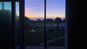 Un balcon offrant une vue sur un coucher de soleil au-dessus d’une zone herbeuse, avec des bâtiments visibles au loin.