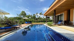 A clear blue pool with tiled edges, surrounded by wooden deck chairs and umbrellas, with a view of lush greenery and a modern building in the background. - Villa Tango with private Pool, Koh Yao Noi  (Ko Yao Noi)