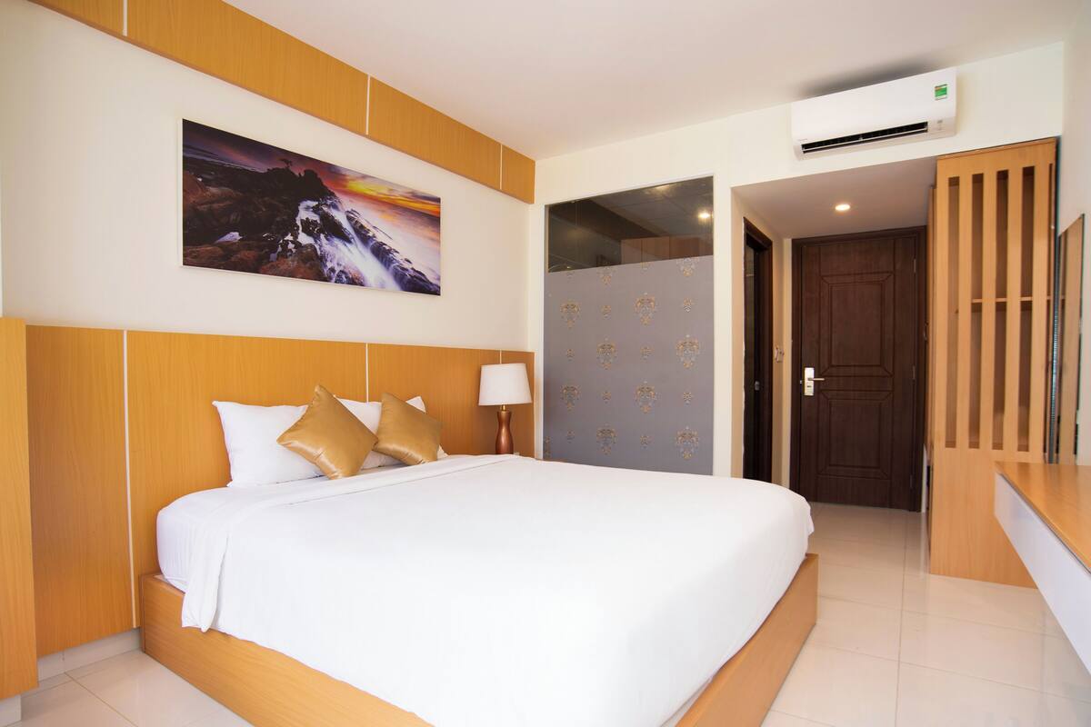 A hotel room with a large bed, wooden headboard, a painting on the wall, and an air conditioning unit.