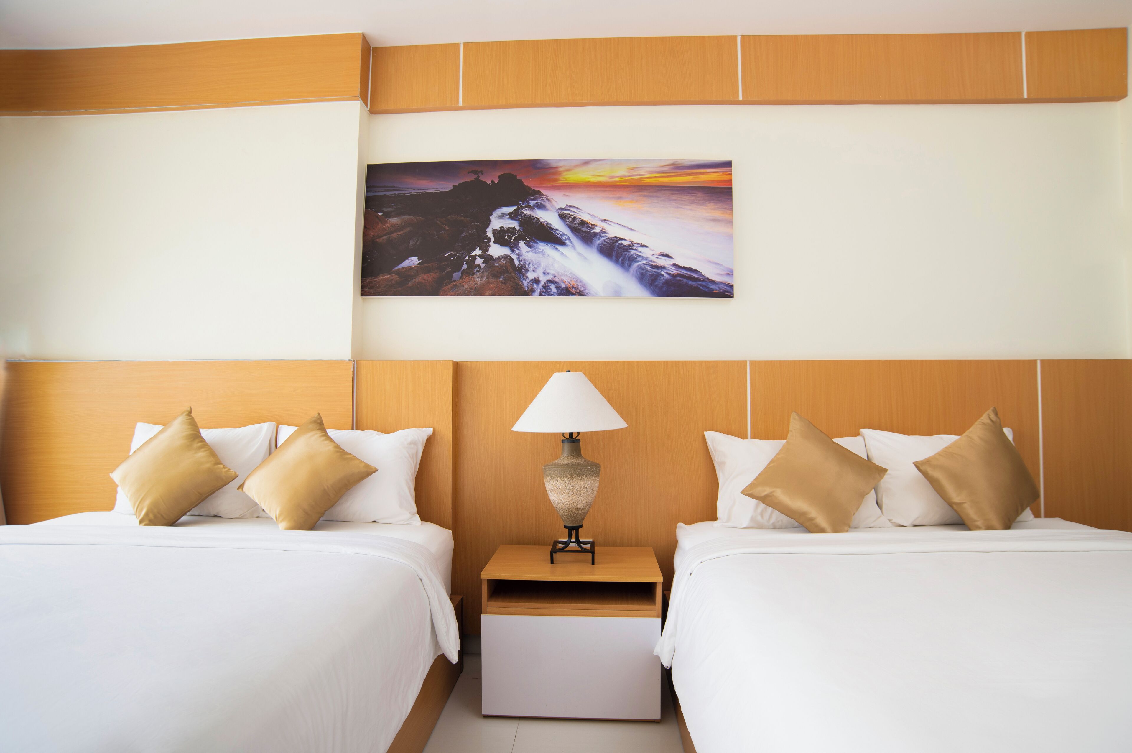 Una camera d'albergo con due letti, un comodino con una lampada e un dipinto raffigurante un tramonto su una costa rocciosa.