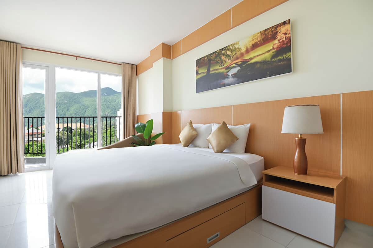 A hotel room with a large bed, a nightstand, a lamp, a painting, and a view of greenery through a balcony.