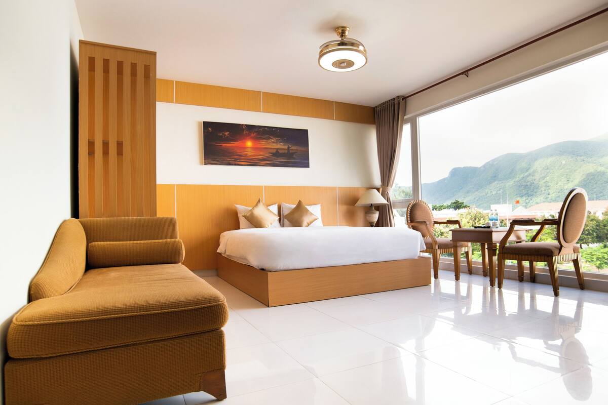 A modern hotel room with a large bed, a sofa, a dining area, and a view of mountains.