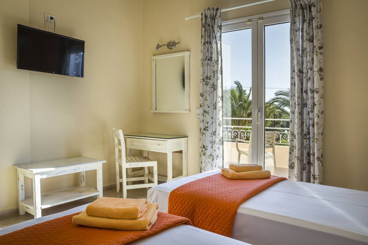 Ein Hotelzimmer mit zwei Betten, einem Schreibtisch, einem Sessel, einem Fernseher und einem Balkon mit Vorhängen.