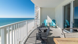 A balcony with a view of the ocean, featuring a white railing, two blue Adirondack chairs, and a wooden table.