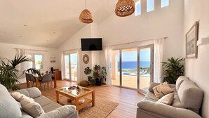 Ein geräumiges Wohnzimmer mit großem Fenster zum Strandblick, einem Essbereich mit Tisch und Stühlen sowie einer gemütlichen Sitzecke mit Sofas und Couchtisch.