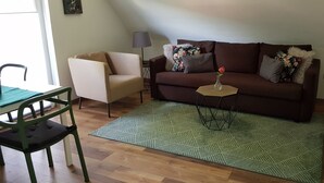 A living room with a brown sofa, a small round table with a vase of flowers, a green patterned rug, a white armchair, and a floor lamp. - Vacation apartment time out! In the middle of the Palatinate Forest. (Erfweiler)