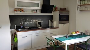 A compact kitchen with white cabinets, a black backsplash, a microwave, a coffee maker, and a small oven. There is a dining table with a green tablecloth and a fruit basket on the side. - Vacation apartment time out! In the middle of the Palatinate Forest. (Erfweiler)