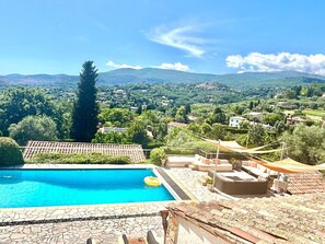 A swimming pool with stone tiles, a lounge area with outdoor furniture, and a view of a village with houses and mountains in the background.