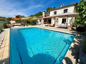 A rectangular swimming pool with clear blue water, surrounded by stone tiles and shaded by umbrellas.