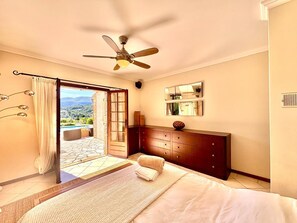 A bedroom with a bed, a wooden dresser, a ceiling fan, a large mirror, and a view of a pool and mountains through an open door.
