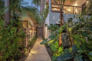 Um caminho ladeado por plantas tropicais exuberantes, levando a um edifício com porta de madeira e escadaria.