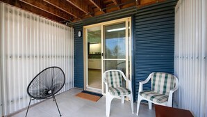 Un patio couvert doté d’un mur en métal ondulé, d’une porte-fenêtre coulissante donnant sur un espace intérieur, et de deux chaises blanches avec des coussins à carreaux.