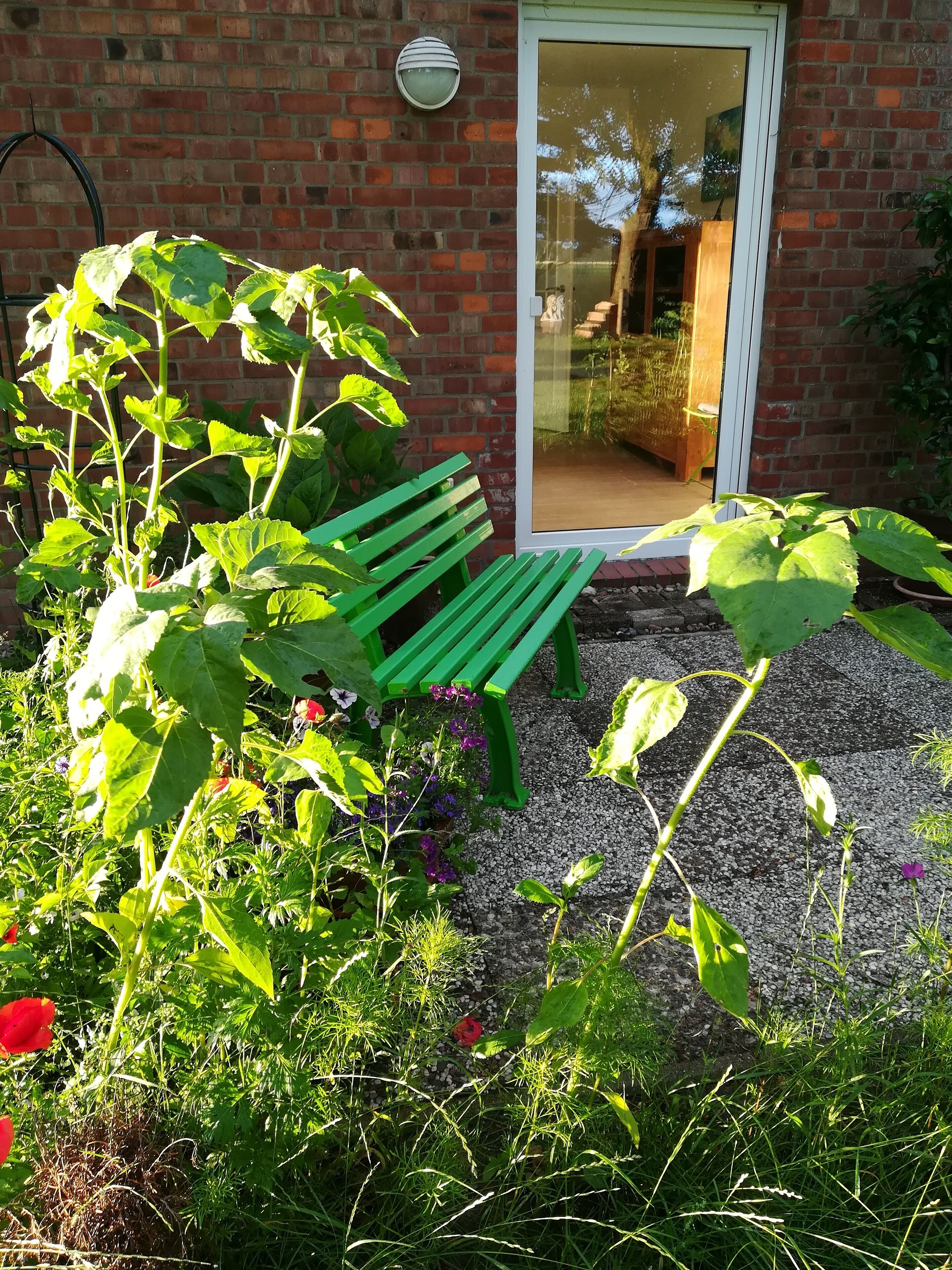 Un jardín con un banco verde, un muro de ladrillo y una puerta que refleja una habitación con una mesa y sillas.