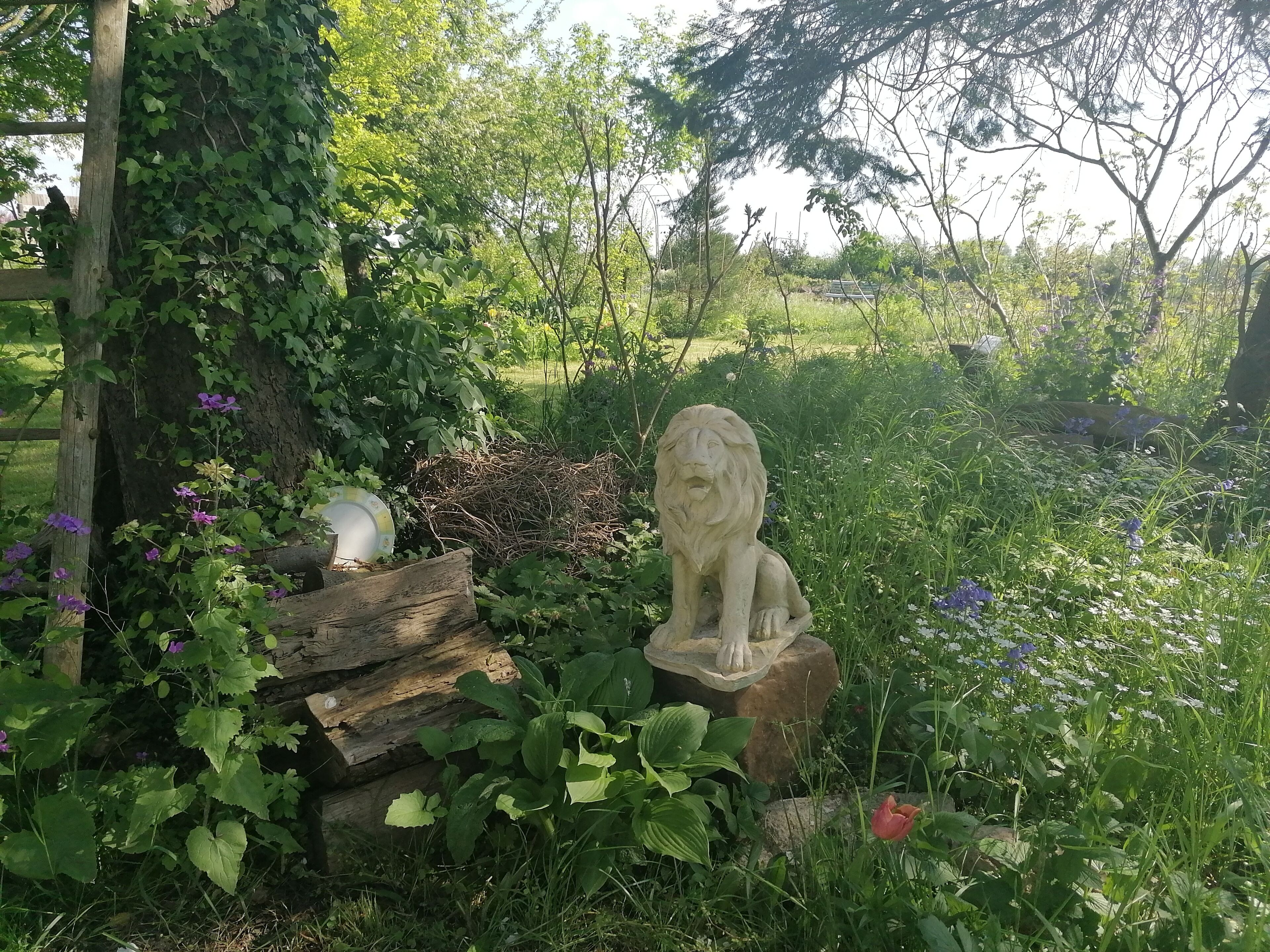 Una estatua de jardín de un león sobre un pedestal de piedra, rodeada de exuberante follaje y flores.