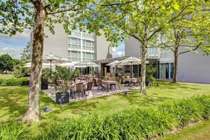 Un patio al aire libre con mesas y sillas, rodeado de árboles y un edificio con grandes ventanales.